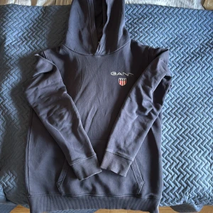 Mörkblå hoodie från GANT - Säljer en stilren mörkblå hoodie från GANT med broderad logga då jag har växt ur den. Tröjan har en klassisk känguruficka och ribbade muddar vid ärmslut och nederkant. Perfekt för en avslappnad stil.