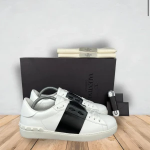 Valentino Garavani sneakers i vitt och svart - Snygga Valentino Garavani sneakers i vitt med en bred svart rem över ovansidan. Skorna har en rund tå och är dekorerade med de ikoniska nitarna på hälen. Perfekta för en stilren look. Hör av er vid funderingar
