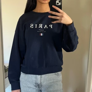 Mörkblå sweatshirt med Paris-tryck - Säljer en snygg mörkblå sweatshirt med texten 'PARIS' och en liten fransk flagga på framsidan. Tröjan har långa ärmar och en bekväm passform. Perfekt för en avslappnad stil.