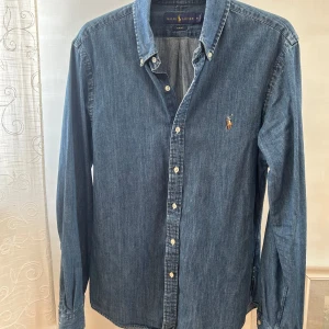 Blå jeansskjorta från Ralph Lauren - Snygg blå jeansskjorta från Ralph Lauren i slim fit. Skjortan har långa ärmar och knappar framtill. Den är broderad med den klassiska logotypen på bröstet. Perfekt för en stilren look. Helt ny utan prislapp.