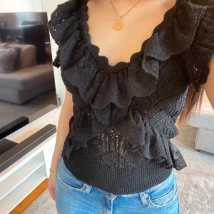 Svart spetsblus med volanger - Snygg svart spetsblus med volanger och korta ärmar. Perfekt för en stilren look med en touch av elegans. Blusen har en v-ringad design och är tillverkad i ett mjukt material som ger en bekväm passform. Storlek L men kan även passa M/S då den är liten i storleken.