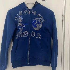 Blå hoodie med tryck från Aim for the Moon - Säljer en blå hoodie med dragkedja och tryck från Aim for the Moon. Hoodien har ett unikt tryck med text och motiv på framsidan. Tillverkad i 100% fransk bomull, vilket gör den mjuk och bekväm. Perfekt för en avslappnad stil. Inte använda så jätte många gånger.