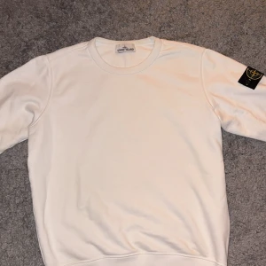 Vit sweatshirt från Stone Island - Säljer en stilren vit sweatshirt från Stone Island. Tröjan har en klassisk rund halsringning och en ikonisk logga på ärmen. Perfekt för en avslappnad och trendig look. Tveka inte på att höra av dig om du har några frågor eller funderingar.