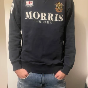 Mörkblå tröja från Morris - Säljer en snygg mörkblå tröja från Morris med broderad logga och text på framsidan. Tröjan har långa ärmar och ribbade muddar vid ärmslut och nederkant. Perfekt för en stilren look! Oanvänd och är stilren med dagens stil! 2 st hål i varsin handled men inuti tröjan inget man ser synligt 