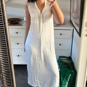 Vit ärmlös klänning - Säljer en stilren vit ärmlös klänning med knappar framtill. Klänningen har en avslappnad passform och är perfekt för varma dagar. Den har en krage och två bröstfickor som ger en snygg detalj. Perfekt för en somrig look! Passar som en M/S. Som referens: jag är 1,76cm!