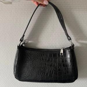 Svart axelväska med krokodilmönster - Snygg svart axelväska med krokodilmönster och dragkedja. Perfekt för att ge din outfit en stilfull touch. Väskan har en bekväm axelrem och är tillverkad i syntetmaterial.