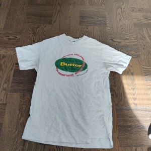 Vit/ljus grå t-shirt från Butter - Säljer en vit t-shirt från Butter med ett färgglatt tryck i grönt och rött på framsidan. T-shirten har en normal passform och korta ärmar. Perfekt för en avslappnad stil.