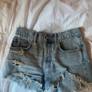 Jeanshorts  - Säljer ett par coola blå jeansshorts från Zara med slitna detaljer och fransar. Perfekta för en avslappnad stil med hög midja och klassisk femficksdesign. Passar perfekt till sommardagar! 🌞