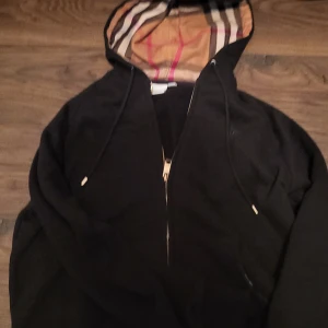 Svart jacka med huva från Burberry - Säljer en stilren svart jacka från Burberry med dragkedja och huva. Insidan av huvan har det klassiska Burberry-rutiga mönstret. Perfekt för en trendig look.
