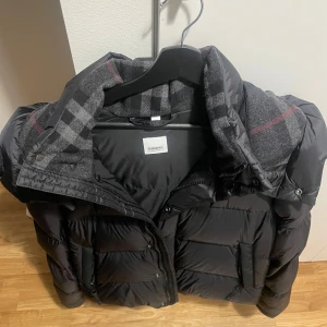 Svart pufferjacka från Burberry - Säljer en snygg svart pufferjacka från Burberry. Jackan har en dragkedja och knappar framtill samt en klassisk rutig insida. Perfekt för kyliga dagar med stil. 🖤