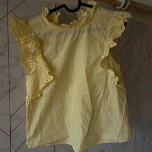 Gul ärmlös blus med spetsdetaljer - Charmig gul blus med spetsdetaljer runt ärmhålen och halsen. Blusen är ärmlös och har en luftig passform, perfekt för varma dagar. Den har en söt och romantisk stil med volanger som ger extra charm.