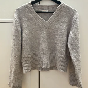 Grå stickad tröja med v-ringning,  - Säljer en mysig grå stickad tröja från ginatricot med v-ringning och ribbade detaljer vid ärmslut och nederkant. Perfekt för kyligare dagar!