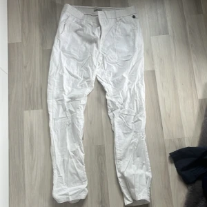 Vita byxor från Blend - Säljer ett par vita byxor från Blend. De har en normal passform och är tillverkade i ett lätt material, perfekt för varmare väder. Byxorna har en elastisk midja och stängs med dragkedja och knapp. Enkla och stilrena för en avslappnad look.