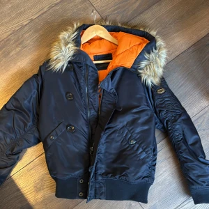 Svart bomberjacka med pälsdetaljer - Snygg svart bomberjacka från Crocker med orange foder och pälsdetaljer på huvan. Jackan har både dragkedja och knappar framtill samt ribbade muddar vid ärmslut. Den har blivit använd ett fåtal gånger