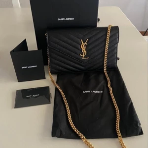 Svart axelväska från Saint Laurent - Elegant svart axelväska från Saint Laurent med quiltad design och guldfärgad kedja. Väskan har en ikonisk YSL-logga på framsidan och är perfekt för att ge en lyxig touch till din outfit. Anledningen varför jag säljer den för så lite är att jag har ont om pengar just nu💕