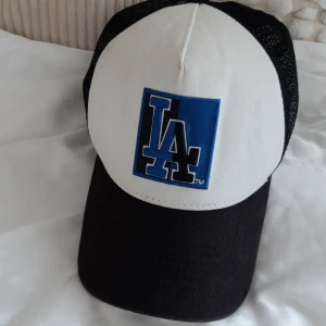 Vit och svart keps med LA-logga - Snygg keps från New Era med LA-logga i blått och svart på framsidan. Kepsen har en vit framsida och svart skärm samt mesh på baksidan för bra ventilation. Perfekt för en avslappnad stil.