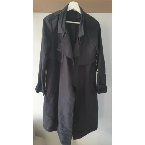 Svart trenchcoat - Snygg svart trenchcoat med långa ärmar och bandet fästes på sidan av midjan i två metallringar. Jackan har en klassisk design med bred krage och en stilren look (och fickor!!) Lite matt i färgen så ser nästan mörkgrå ut i visst ljus. 57% modal, 43% polyester. 