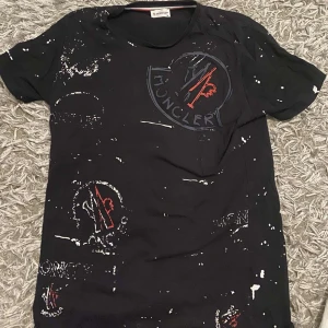 Svart t-shirt från Moncler - Säljer en svart t-shirt från Moncler med coolt stänkmönster i vitt. Den har korta ärmar och en normal passform. Perfekt för en avslappnad stil eller fest. Märkesloggan syns tydligt på framsidan. Passar bra till jeans eller shorts!