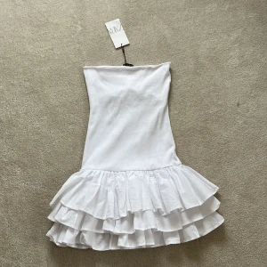 Vit axelbandslös klänning från Zara - Säljer en stilren vit axelbandslös klänning från Zara. Klänningen har en volangdetalj vid fållen som ger en lekfull touch. Perfekt för sommarens alla tillfällen. Helt oanvänd. OBS!! Väldigt liten i storleken så skulle säga att den passar S & Xs , är själv Xs vanligtvis men den passar perfekt 