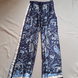 Mönstrade finbyxor i satin - Mina favorit-finbyxor med paisleymönster i blått och beige. Byxorna har en utsvängd passform och är tillverkade i ett mjukt och lätt material (satin) som glänser i visst ljus. Dragkedja bak och vida ben 💙 på bild 5 syns en tråd som ser ut som den dragits på nära håll, vilket inte syns när man har de på sig 