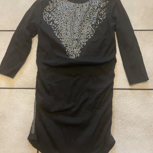 Svart klänning med paljetter storlek XS/S - Snygg svart klänning med glittriga paljetter i ett triangulärt mönster framtill. Klänningen har trekvartsärmar och en smickrande passform. Perfekt för en kväll ute!