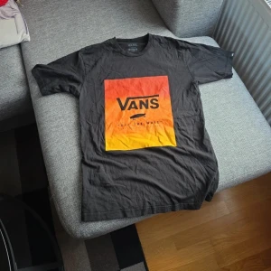 Svart t-shirt från Vans - Säljer en svart t-shirt från Vans med ett coolt tryck i orange och gult på framsidan. T-shirten har korta ärmar och är perfekt för en avslappnad stil. Den är gjord i bomull och har en klassisk passform. Perfekt för alla som älskar streetwear! 🛹