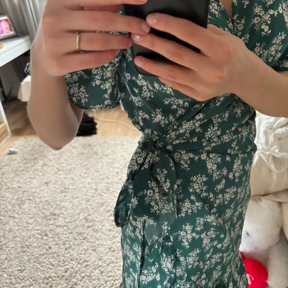 Säljer en söt grön omlottklänning från Aachoe med vita blommor. Klänningen har korta ärmar och en knytning i midjan som ger en fin passform. Perfekt för sommarens alla tillfällen! 🌸Storleken är S men eftersom den har knytningen kan man justera den så skulle säga att den passar XS-M🥰. Mekot.