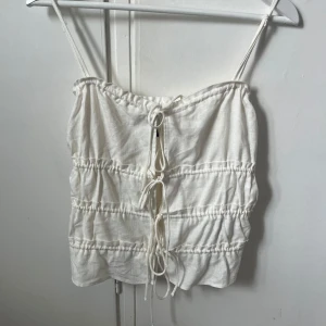 Vit linnetopp med knytning - Säljer en söt vit linnetopp med smala axelband och knytning framtill. Toppen har en luftig och avslappnad passform med dekorativa rynkdetaljer. Perfekt för varma sommardagar! ❤️