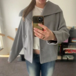 Grå kappa med knappar - Snygg grå kappa med stora knappar och bred krage. Jackan har en klassisk design med en något lös passform och långa ärmar. Perfekt för kyligare dagar.