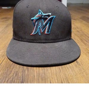 Svart keps med broderad logotyp - Snygg svart keps från New Era med en broderad logotyp i blått och rött framtill. Kepsen har en klassisk design med en rak skärm och är perfekt den är 58,7 cm de står mitt efternamn på vänster sida men priset är inte hugget I sten skriv om du har några frågor eller funderingar 