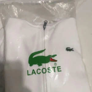 Vit hoodie från Lacoste - Säljer en stilren vit hoodie från Lacoste med dragkedja och den klassiska krokodilloggan på bröstet. Tröjan har en justerbar huva och praktiska fickor framtill. Perfekt för en avslappnad look. Kan även diskuteras om pris vid snabb affär. Storlek M funkar på S. Aldrig använt för den är för liten