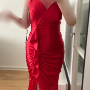 Röd långklänning med volangdetalj - Snygg röd långklänning med v-ringning och volangdetalj framtill.                                      Helt ny aldrig använd, prislapp kvar Slits framtill Storlek XS, kan passa S  Pris kan diskuteras. Fler bilder kan skickas vid intresse. Hör av dig om du har frågor