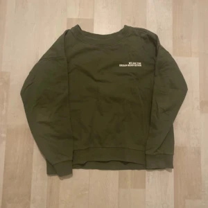 Olivgrön sweatshirt  - Olivgrön sweatshirt med rund halsringning och tryck i vitt på bröstet. Tröjan har en avslappnad passform och långa ärmar, perfekt för en chill och stilren look. Tillverkad i mjuk bomull för extra komfort.