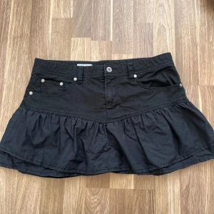 Svart jeans volangkjol från Clockhouse  - Supersnygg svart kortkjol från Clockhouse med volang nertill och klassiska jeansdetaljer som fickor och bälteshällor. Kjolen är gjord i 100% bomull och har en cool, lite utställd form som ger en trendig vibe. Den sitter mer som en storlek M!