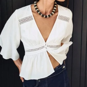 Vit blus med v-ringning från Zara - Superfin vit blus från Zara med djup v-ringning och trekvartsärmar. Blusen har dekorativa knappar framtill, broderade detaljer och lätt volang nedtill. Luftigt bomullstyg som passar perfekt till jeans eller kjol.