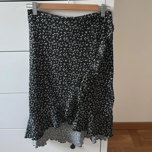 Kjol Gina Tricot Young  - Svart kjol från Gina Tricot Young med små vita blommor över hela tyget. Kjolen har volangkant nertill och en lätt vågig form. Stl 158/164.