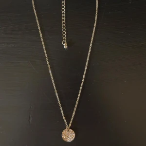 Guldigt halsband med rund berlock - Snyggt halsband i guldton med tunn kedja och en rund berlock med ingraverat mönster. Perfekt för att ge din outfit en extra touch av stil. Justerbar längd med förlängningskedja. Mycket bra skick