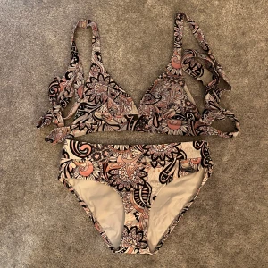 Mönstrad bikini från Odd Molly - Supersnygg bikini från Odd Molly med blommigt mönster i rosa, beige, svart och vitt. Överdelen har knytband vid axlarna och byxan har en liten metallogo framtill. Perfekt för stranden eller poolen i sommar!