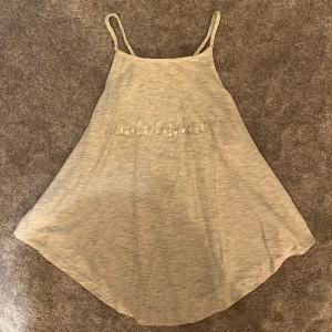 Ljusgrå linnetopp med text - Superskön ljusgrå linnetopp med tunna, flätade axelband och avslappnad passform. Tryckt text framtill: 'and so the adventure begins'. Perfekt för varma dagar eller som ett lager under en hoodie.