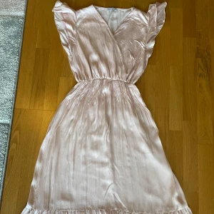Ljusrosa omlottklänning i satin - Superfin ljusrosa långklänning i glansigt satinliknande material. Klänningen har omlottliv, volangdetaljer vid axlar och nederkant samt resår i midjan för en snygg passform. Perfekt för sommarens alla tillfällen! S/M
