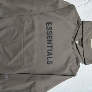 Grön Essentials hoodie - Säljer en grön hoodie från Essentials Fear of God med stor svart logga på bröstet och mindre tryck på ärmen. Klassisk känguruficka, huva och ribbade muddar. Perfekt för dig som gillar streetwear och enkel stil.