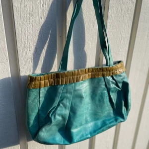 Turkos axelväska i skinn med detaljer - Snygg turkos axelväska i mjukt skinn med unika veckade detaljer i beige upptill. Väskan har långa axelband och rymligt innerfack med dragkedja. Perfekt för dig som vill sticka ut med färg och cool design.