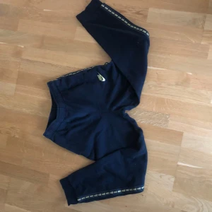 Svarta Nike mjukisbyxor  - Snygga svarta mjukisbyxor från Nike med elastisk midja och guldig Nike-logga på benet. Sidorna har en guldig rand med Nike-tryck längs hela benet. Perfekta för chill eller träning, riktigt sköna och stilrena.