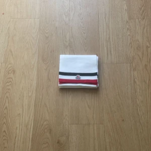 Vit halsduk från Moncler med ränder - Vit halsduk från Moncler med klassiska svarta och röda ränder samt broderad logotyp framtill. Halsduken är stickad och har en clean, stilren look som passar perfekt till vinterjackan.