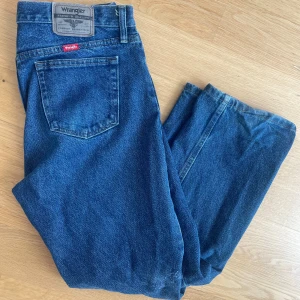 Blå jeans från Wrangler straight fit - Klassiska blå jeans från Wrangler med rak passform och femficksdesign. Jeansen har en mid-midja. Snyggt Wrangler-märke bak i midjan och röd etikett vid bakfickan. Perfekta för en avslappnad och tidlös look.