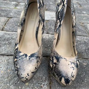 Ormskinnsmönstrade pumps från Topshop - Snygga pumps med ormskinnsmönster i beige och svart från Topshop. Skorna har spetsig tå, hög klack och plattform framtill. Tillverkade i syntetmaterial med en cool textur som ger extra edge till outfiten.