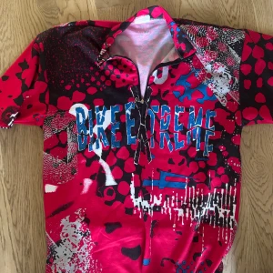 Färgstark pikétröja BIKE XTREME - Säljer en riktigt cool pikétröja från BIKE XTREME med galet färgstarkt mönster i rött, svart, blått och vitt. Tröjan har korta ärmar, halv dragkedja och tryckt text framtill. Perfekt för dig som vill sticka ut med en unik stil.