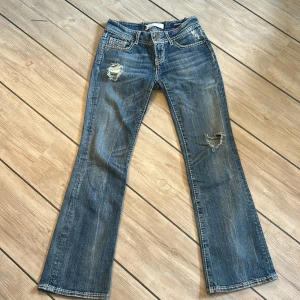 Blå bootcut jeans från Vigoss - Snygga blå bootcut jeans från Vigoss med slitningar och hål på benen för en cool, avslappnad look. Tydliga kontrastsömmar och broderade bakfickor ger extra detaljer, även dubbla knappar i midjan.