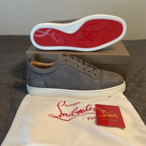 Grå mockasneakers från Christian Louboutin - Snygga grå sneakers i mocka från Christian Louboutin med klassisk vit sula och ikonisk röd undersida. Skorna har rund tå, snörning och diskreta detaljer. Insidan är fodrad i beige läder för extra komfort. Perfekta för dig som gillar stilrena och exklusiva sneakers.⚠️ Äkthet kan ej granskas där av priset 