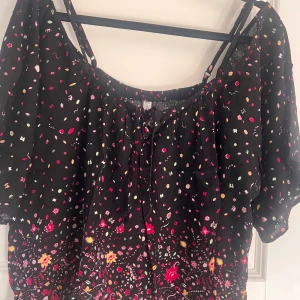 Svart blommig blus från FB Sister - Supersöt svart blus från FB Sister med färgglatt blommönster i rosa, rött, gult och grönt. Modellen har korta ärmar, smockad halsringning med snörning och tunna axelband. Luftigt tyg i 100% viskos, perfekt för en chill och trendig look.